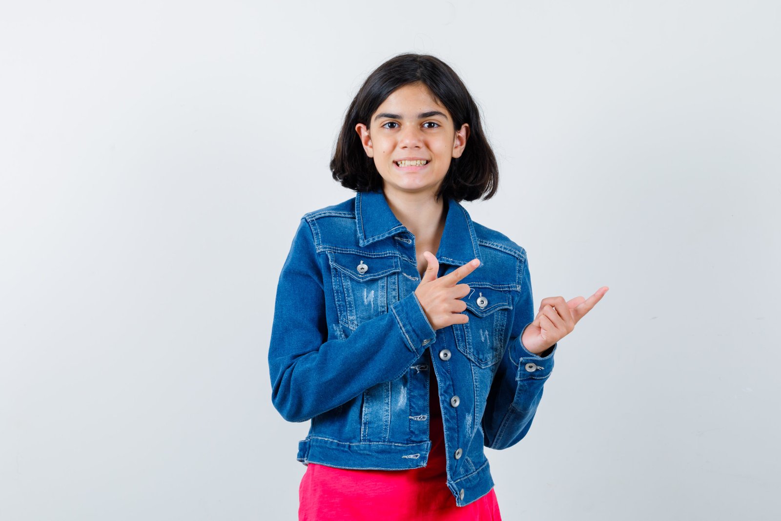 young girl pointing right with index fingers red t shirt jean jacket looking happy front view scaled